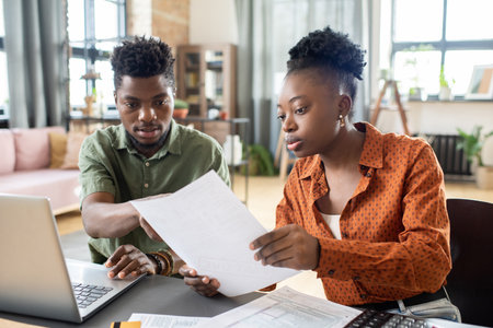 Young blackwoman showing document to husband while discussing tax papers with him at homeの写真素材