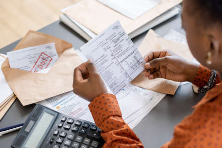 Over shoulder view of blackwoman sitting at table with bills and calculator and reading receiptの写真素材