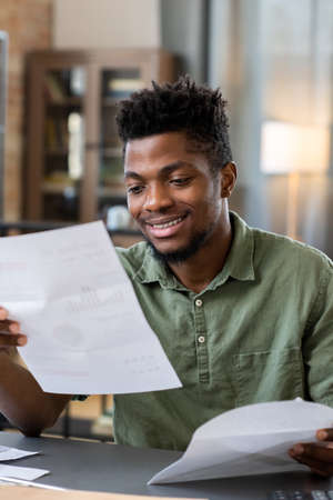 Smiling young Blackman with beard sitting at table in living room and receiving letter from bankの写真素材