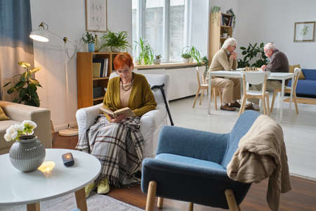 Elderly woman sitting in armchair in the room and reading a book with men playing chess in the backgroundの写真素材