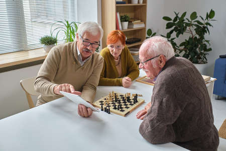 Senior man pointing at digital tablet and explaining the rules of game to his friends while they sitting at the table with chess boardの写真素材