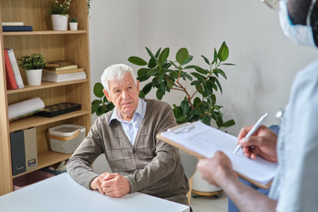 Elderly man sitting at the table and listening the prescription of his doctor while he making notes in medical cardの写真素材