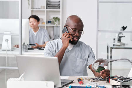 Young African repairman speaking on mobile phone to client in front of laptop in workshopの写真素材