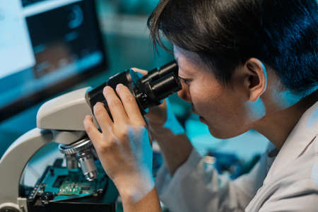 Young Asian technician looking in microscope while studying microchip characteristics in laboratoryの写真素材