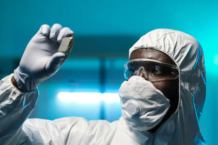 Gloved African scientist in protective coveralls, eyeglasses and respirator holding microchip while working in laboratoryの写真素材