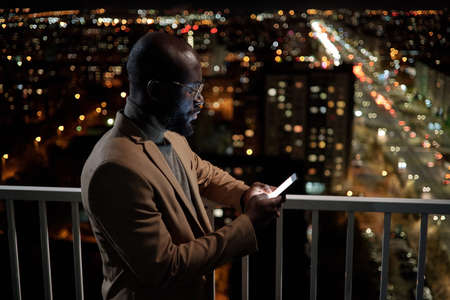 African businessman in formalwear looking through contacts in mobile phone while standing on balcony...