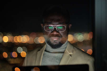 Serious African businessman in eyeglasses looking at computer screen while working late in the nightの写真素材
