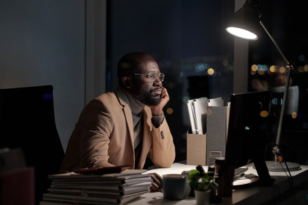African businessman sirring by desk in front of computer monitor in office while working late at nightの写真素材