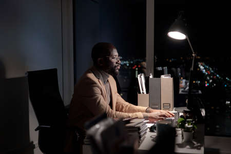 Busy young African man typing on computer keyboard and looking at screen while working late in...