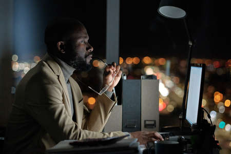 Serious businessman in suit looking at computer screen while sitting by workplace in office at nightの写真素材