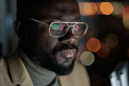 Young African businessman in eyeglasses concentrating on network while working late at night in...