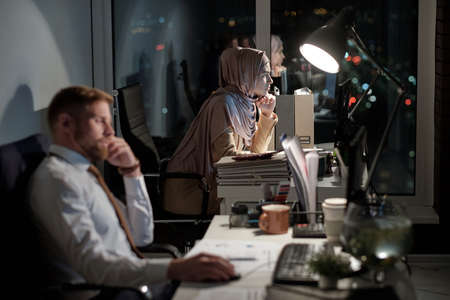 Young serious Muslim businesswoman looking at computer screen while sitting by workplace in office at nightの写真素材