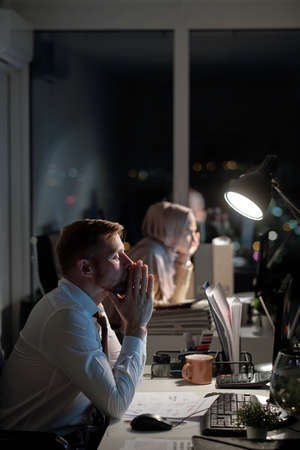 Tired businessman with his hands put together by face sitting in front of computer monitor against...