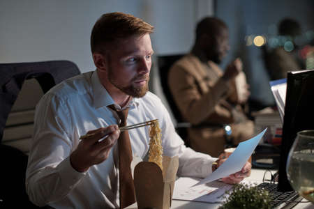 Busy white collar worker with paper eating chinese wok and looking at computer screen in office at nightの写真素材