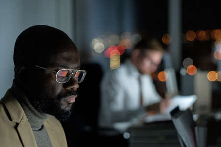 Young serious businessman in eyeglasses looking at computer screen while working late in the evening...