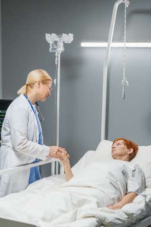 Mature female doctor holding hand of her patient and talking to her while she lying on hospital bedの写真素材