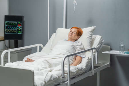 Elderly sick woman with sad expression lying in hospital bed after operationの写真素材