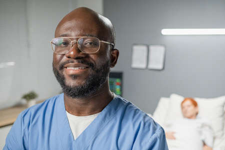 Close-up of African surgeon in eyeglasses smiling at camera while standing at hospital ward with...