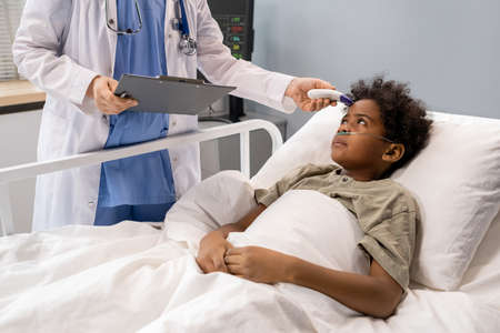 African little boy lying in bed and looking at doctor while she examining the temperature with medical equipment at hospital wardの写真素材