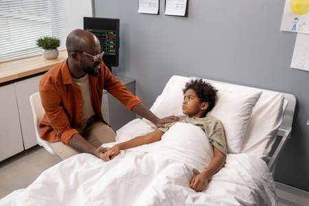 African father talking to his little son while he lying in bed, he visiting him at hospital wardの写真素材