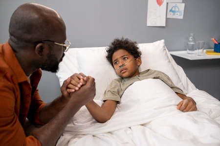 African father holding hand of his son and talking to him during his visit at hospital wardの写真素材