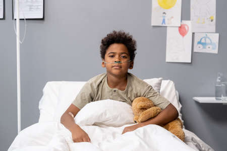 Portrait of African little boy looking at camera while sitting in bed with teddy bear during his treatment at hospitalの写真素材