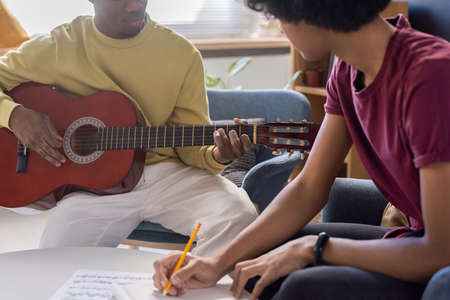 Young biracial guy making notes and looking at music teacher playing guitar during lesson in home environmentの写真素材