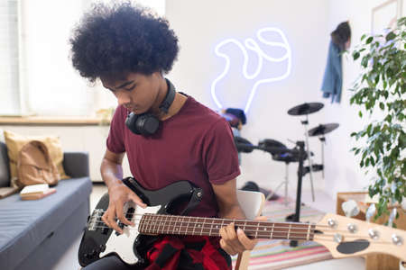 Young serious man with headphones around his neck playing electric guitar while sitting in home...