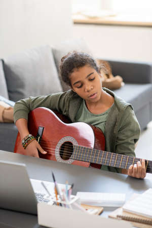 Cute preteenage girl with acoustic guitar playing in front of laptop during online lesson in home environmentの写真素材