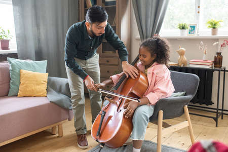 Young man bending over cute diligent schoolgirl with cello while teaching her how to play musical instrumentの写真素材