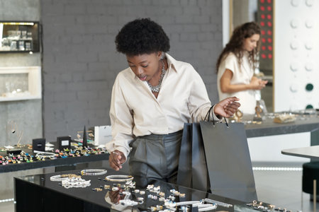 Young African female shopper with paperbags choosing new necklace on large display while looking at jewelry assortmentの写真素材