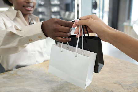 Hands of two young intercultural females holding paperbags over counter after buyer has paid for the...