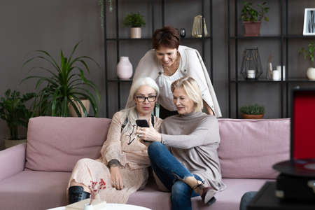 Happy mature woman showing something on her mobile phone to her friends while they sitting on sofa...