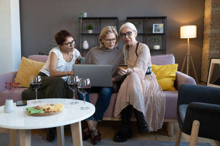Mature women sitting on sofa with laptop using credit card to do shopping online in the living roomの写真素材