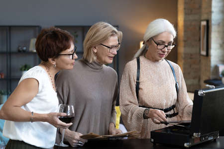 Mature women in eyeglasses listening to music on old player during their home party with wineの写真素材