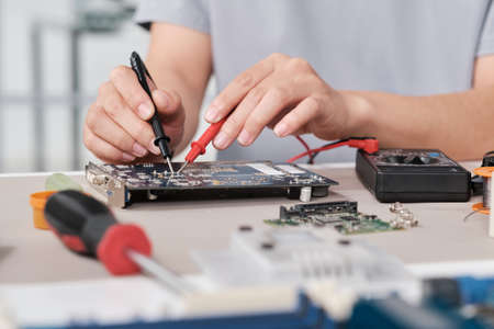 Hands of young female engineer holding soldering iron tweezers close to tiny detail of circuit board...