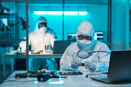 Young female scientist in protective workwear looking at microchip through magnifying glass against...