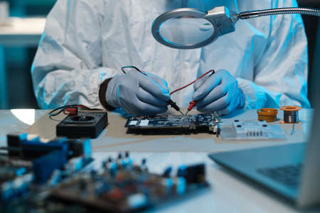 Gloved technician or repairman soldering details of motherboard with tweezers while sitting by...