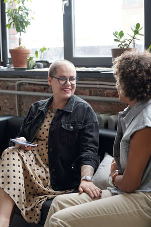 Happy young Caucasian businesswoman with mobile phone talking to her biracial colleague at working...