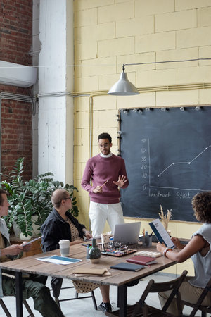 Young confident businessman explaining financial graph to colleagues at seminar while standing by...
