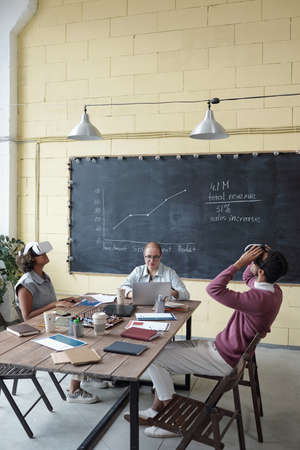 Three young contemporary colleagues sitting by table in office while two of them making virtual...