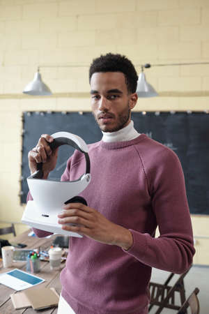 Young confident businessman with vr headset in hands looking at camera while standing against blackboard and workplaceの写真素材