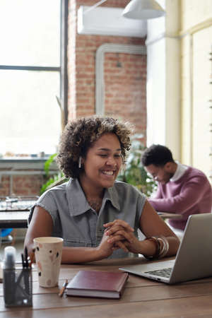 Happy young businesswoman with earphones watching online video while sitting by table in front of...