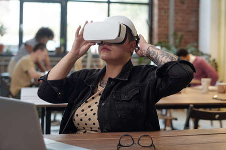 Young female office worker in vr headset watching virtual presentation while sitting by workplace in...