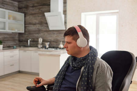 Serious man in headphones listening to music while moving in wheelchair along spacious living-room...