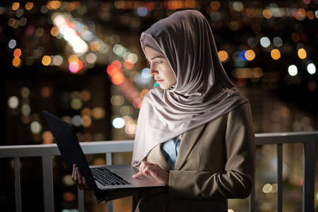 Serious young businesswoman with laptop looking at its screen while networking against urban environment at nightの写真素材