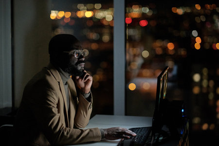 Young serious African businessman looking through online data while sitting in front of computer monitor in officeの写真素材