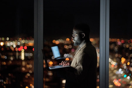 Young businessman with laptop standing against large window in office and analyzing online data late at nightの写真素材