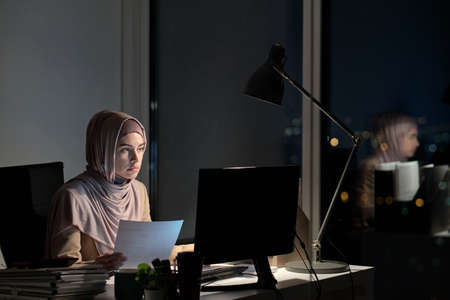 Young Muslim female in hijab holding paper and looking at computer screen while sitting by desk and...