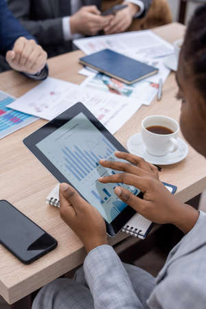 Hand of young African businesswoman scrolling in tablet with charts and diagram on screen while sitting by tableの写真素材
